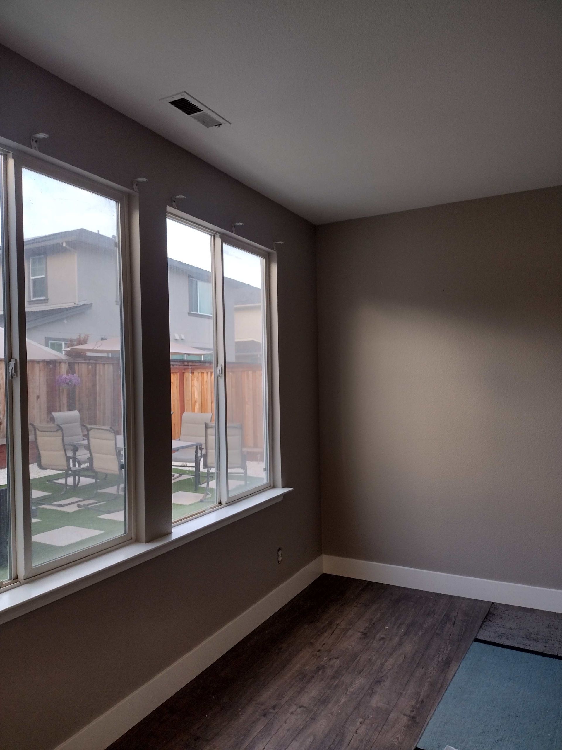 A bright room with two large windows overlooking a backyard, featuring grey walls and dark wood-look flooring.