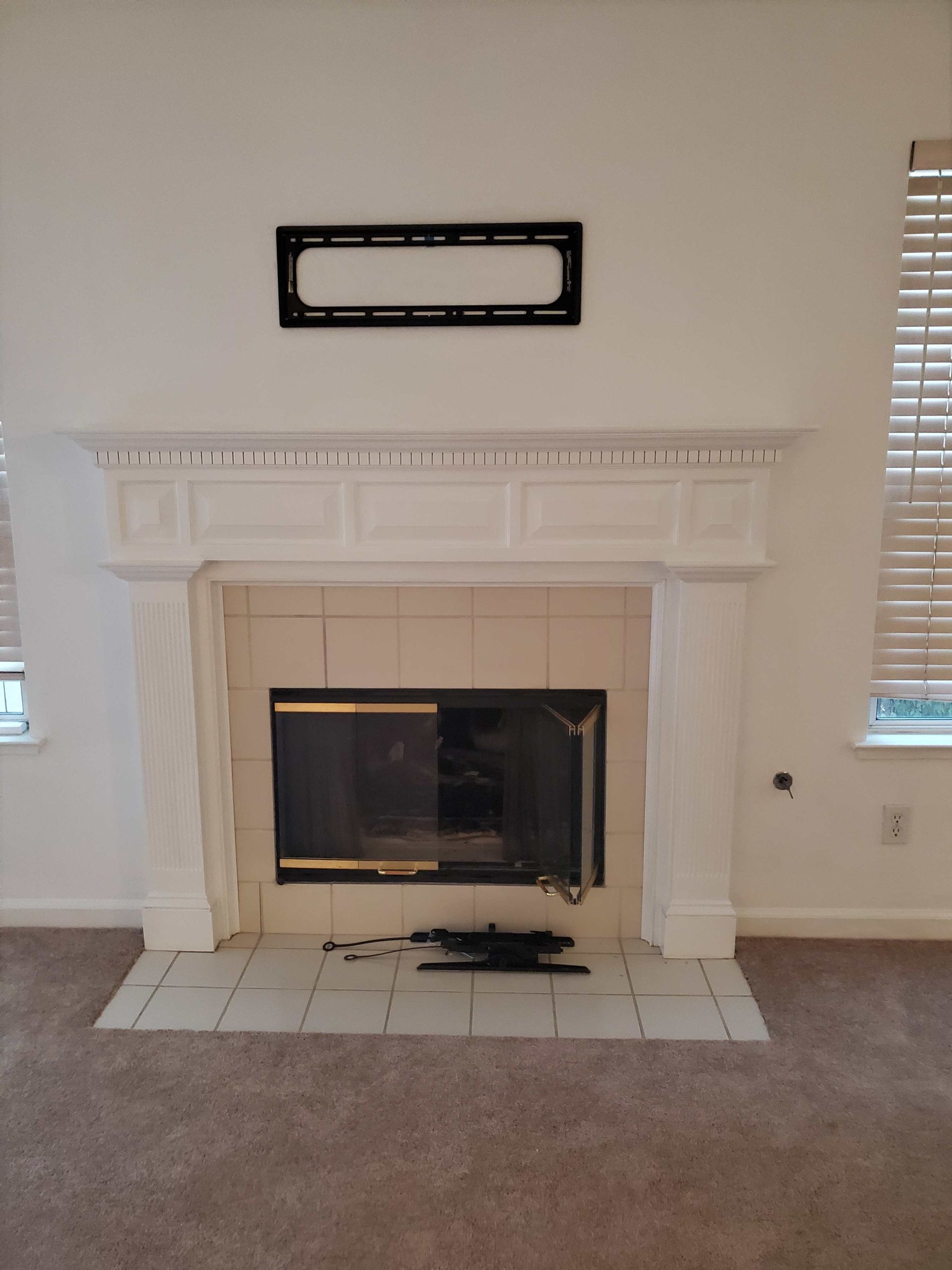 Living room fireplace with white mantel, beige walls, carpet, and a wall-mounted TV above.
