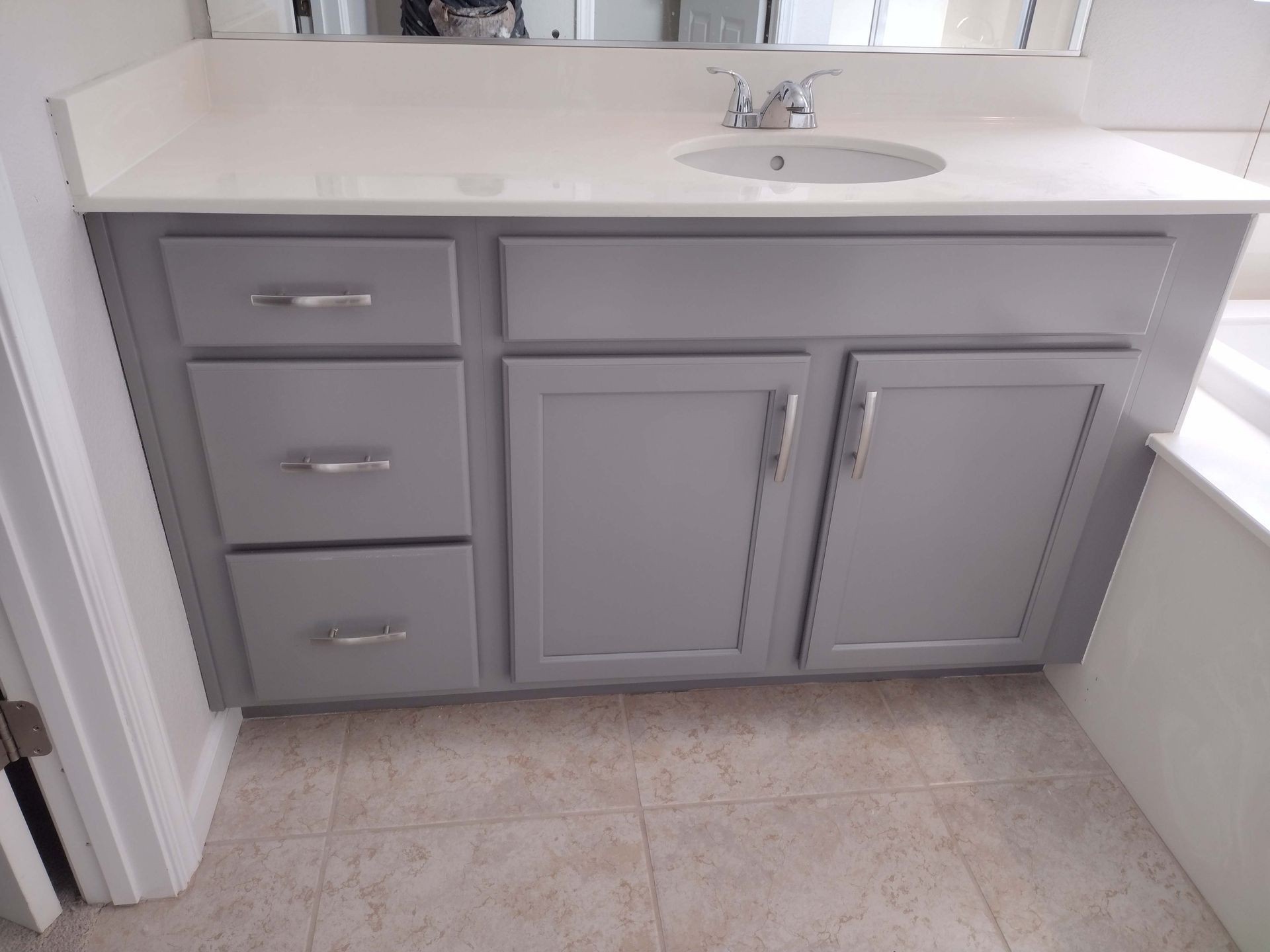Gray bathroom vanity with white countertop, sink, and three drawers beside cabinet doors on a tiled floor