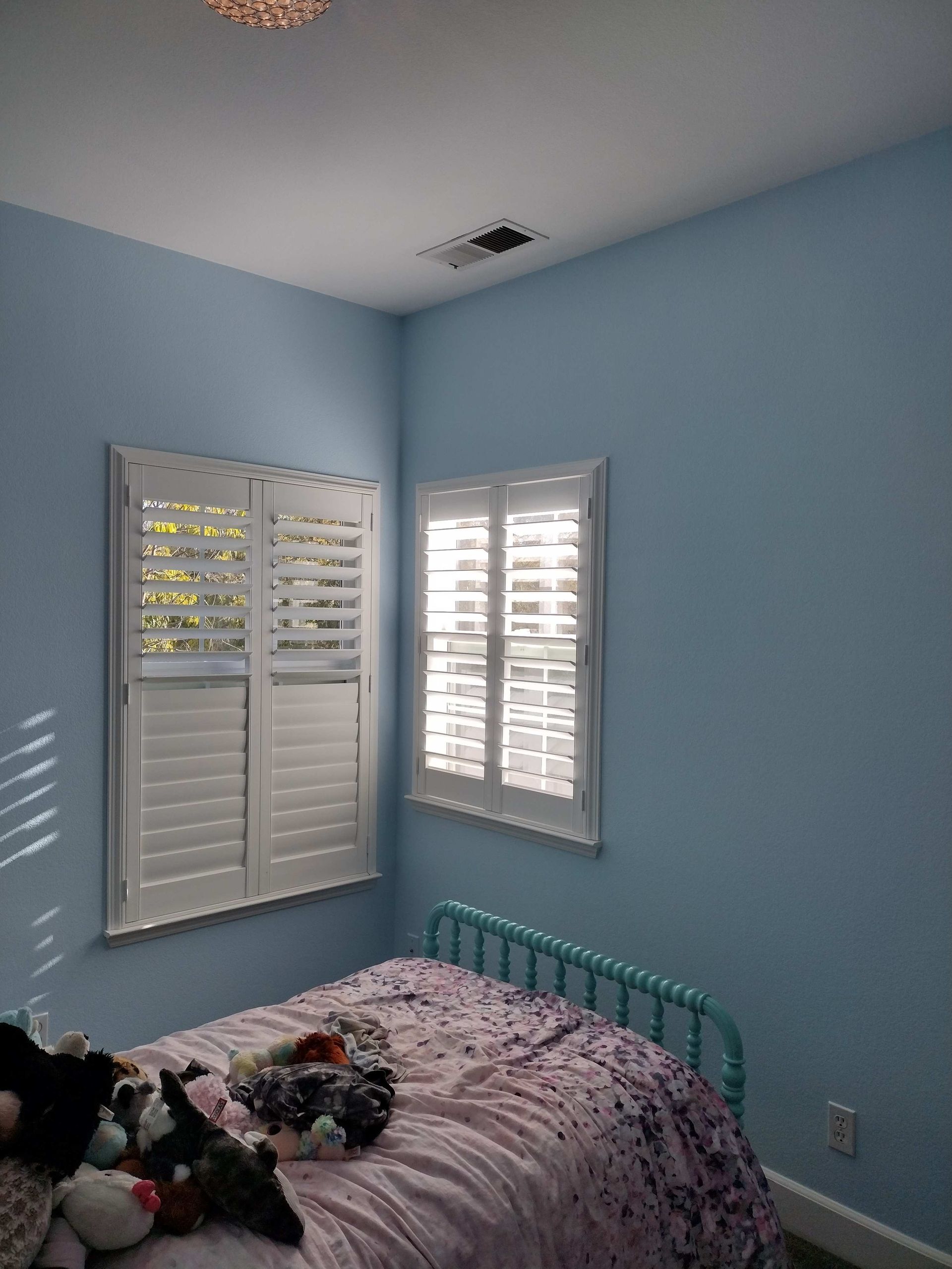 Light blue bedroom with white shutters, teal bed frame, and a floral blanket near two windows.