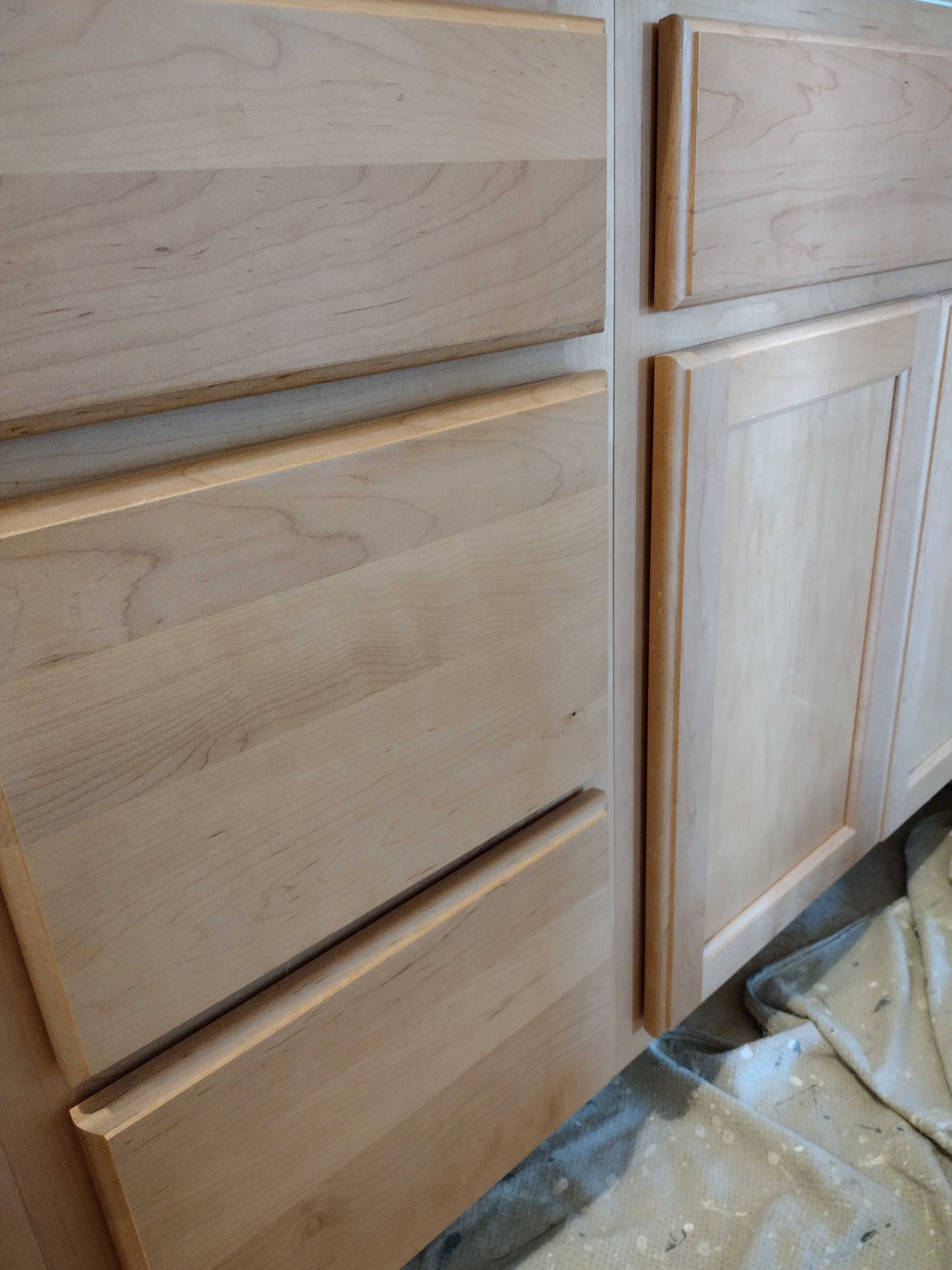 Unfinished wooden cabinet doors with light stain and panel details, partly covered by plastic drop cloth