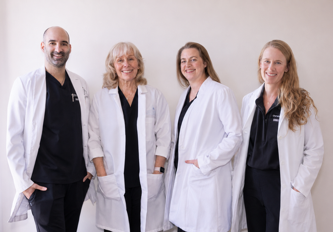 Three smiling medical professionals in white lab coats and black scrubs standing side-by-side against a white wall.