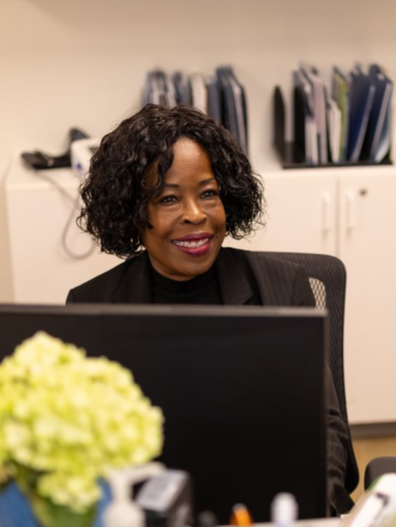Woman smiling at an office desk with a computer and yellow flowers in the foreground.