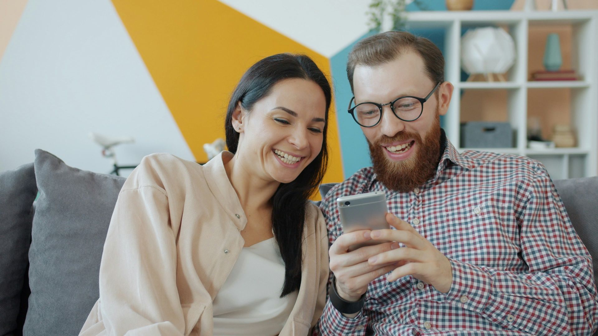 Couple laughing while looking at a phone screen on a gray couch; colorful wall in background.