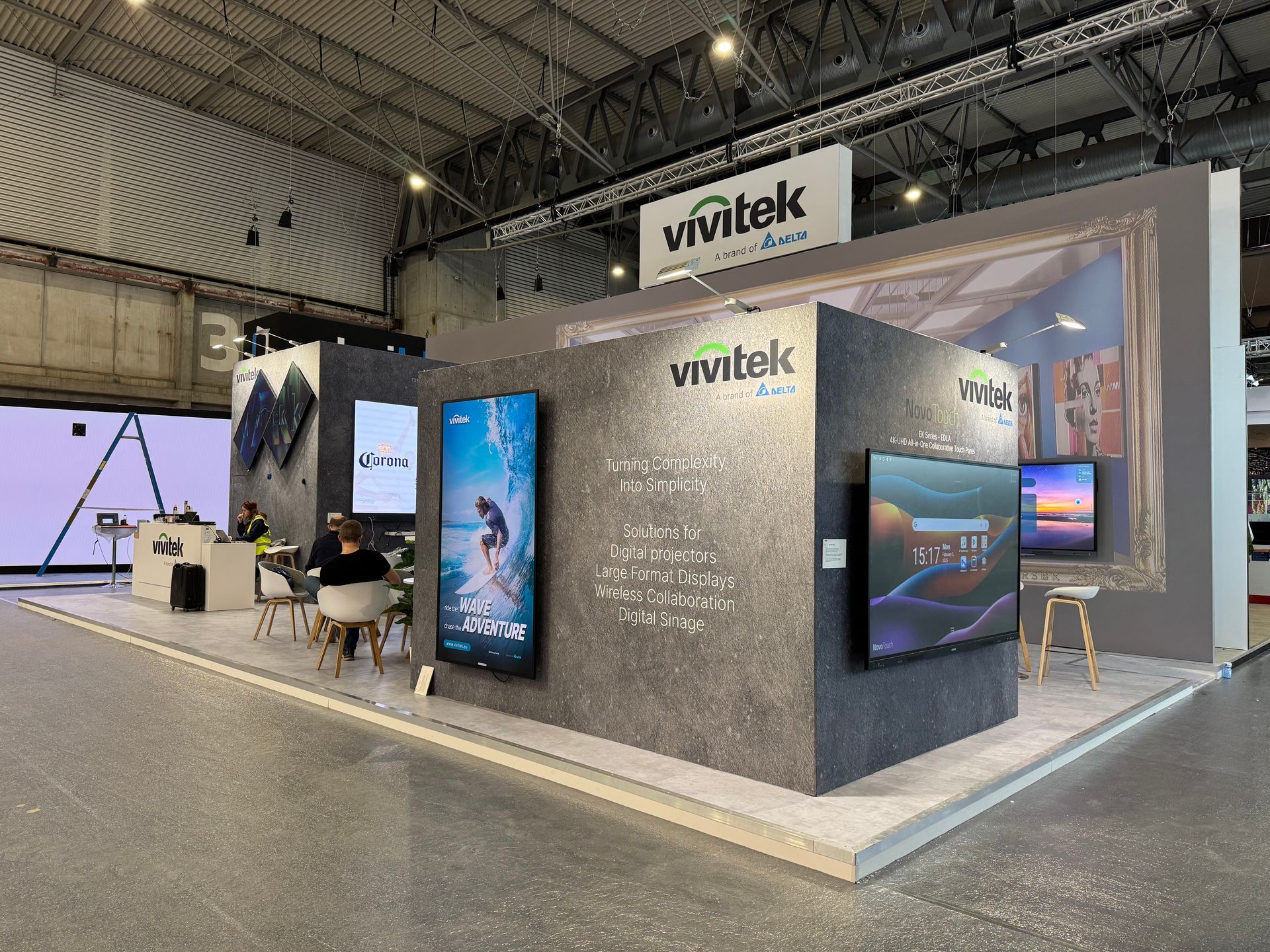 A bright trade show booth featuring Delta and Vivitek displays, wall-mounted screens, and a small table with white chairs.