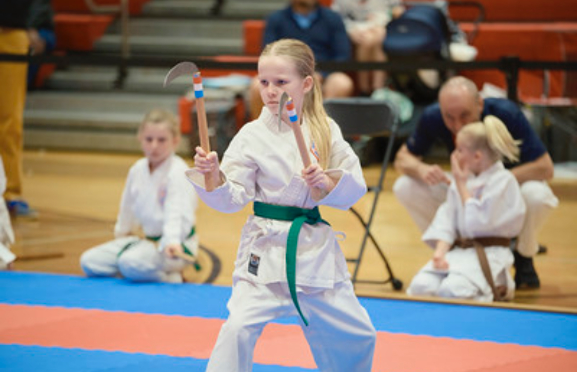 A little girl in a karate uniform is holding a wooden stick.