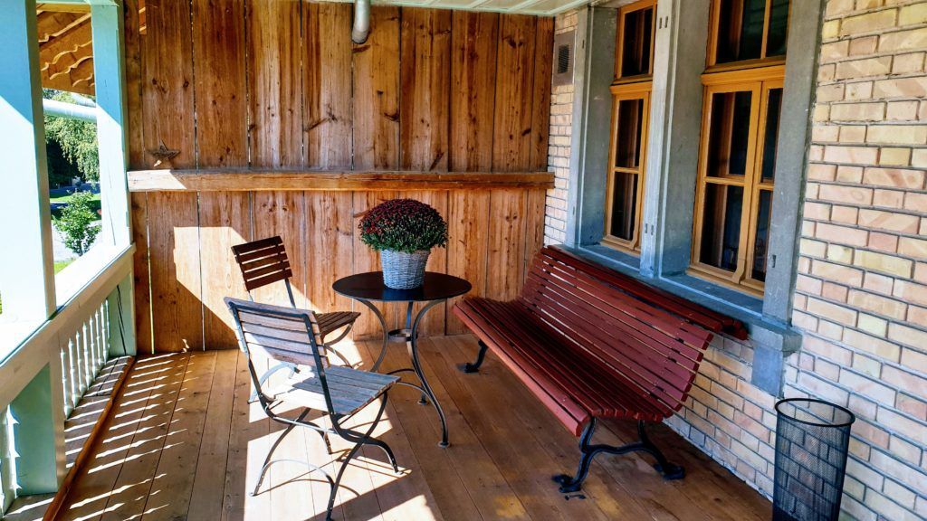 Holzveranda mit Bank, Tisch, Stühlen und Backsteinmauer. Auf dem Tisch steht ein Blumentopf.