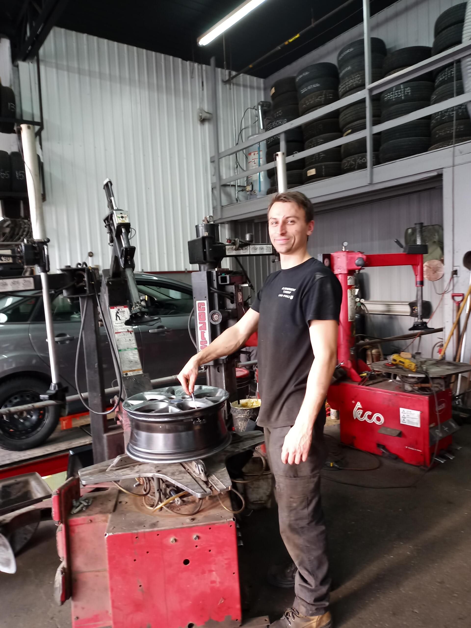 Un homme se tient debout dans un garage automobile, à côté d'une machine à pneus, avec des pneus empilés et des outils en arrière-plan.