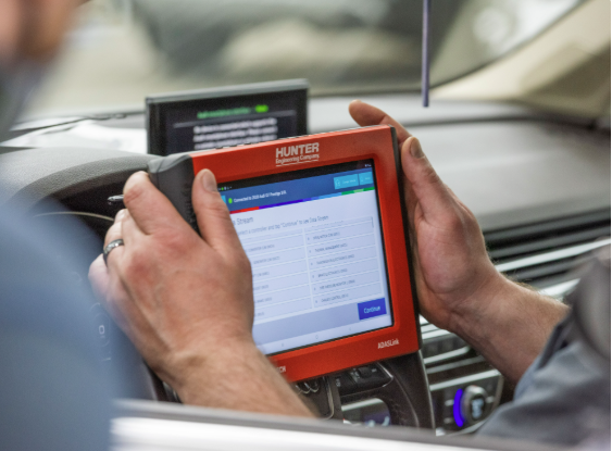 Des mains tiennent une tablette à coque rouge à l'intérieur d'une voiture, affichant un écran de diagnostic.