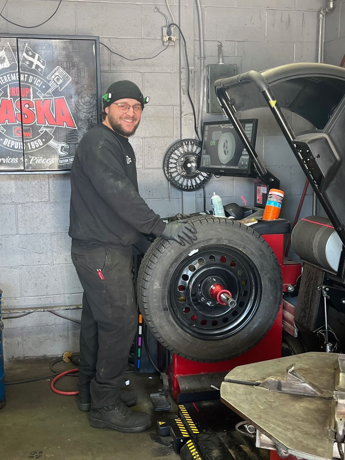Un mécanicien, souriant, tient un pneu à côté d'un pick-up tout-terrain surélevé dans un garage.