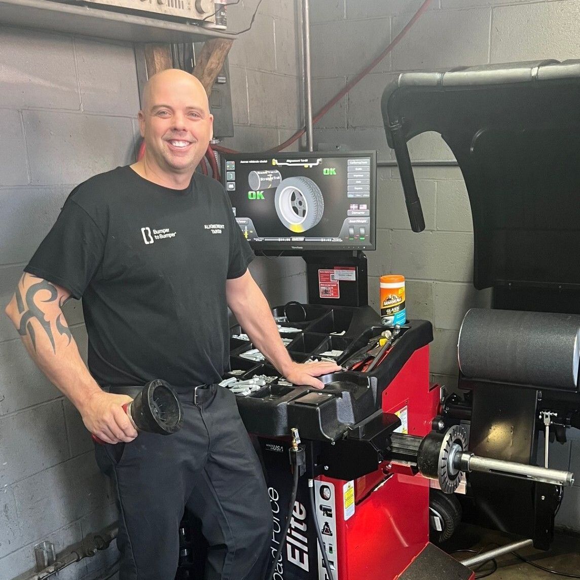 Un homme sourit à côté d'une machine à équilibrer les pneus dans un atelier, avec un pneu de voiture et un écran derrière lui.