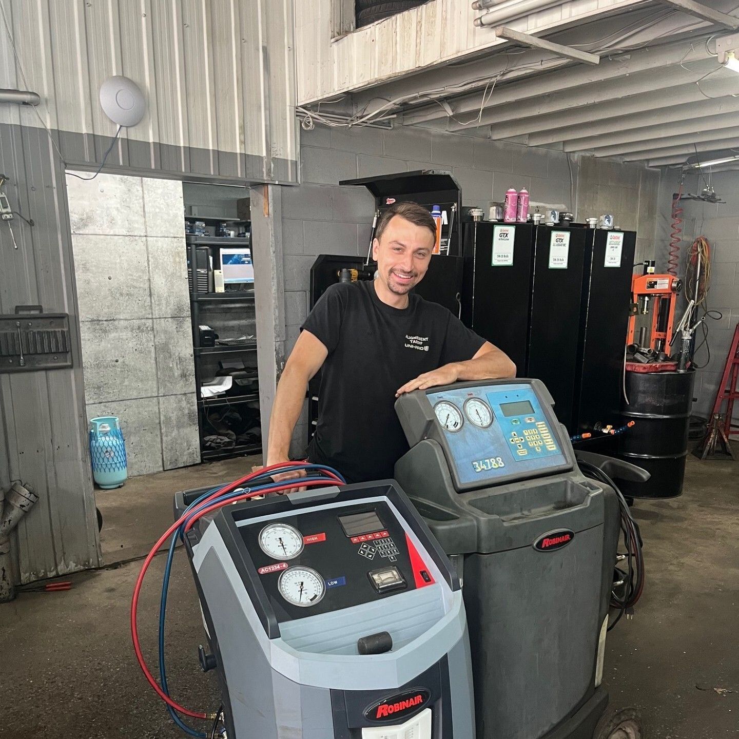 Un homme se tient dans un garage, debout derrière deux machines de réparation de pneus, avec des outils et du matériel en arrière-plan.