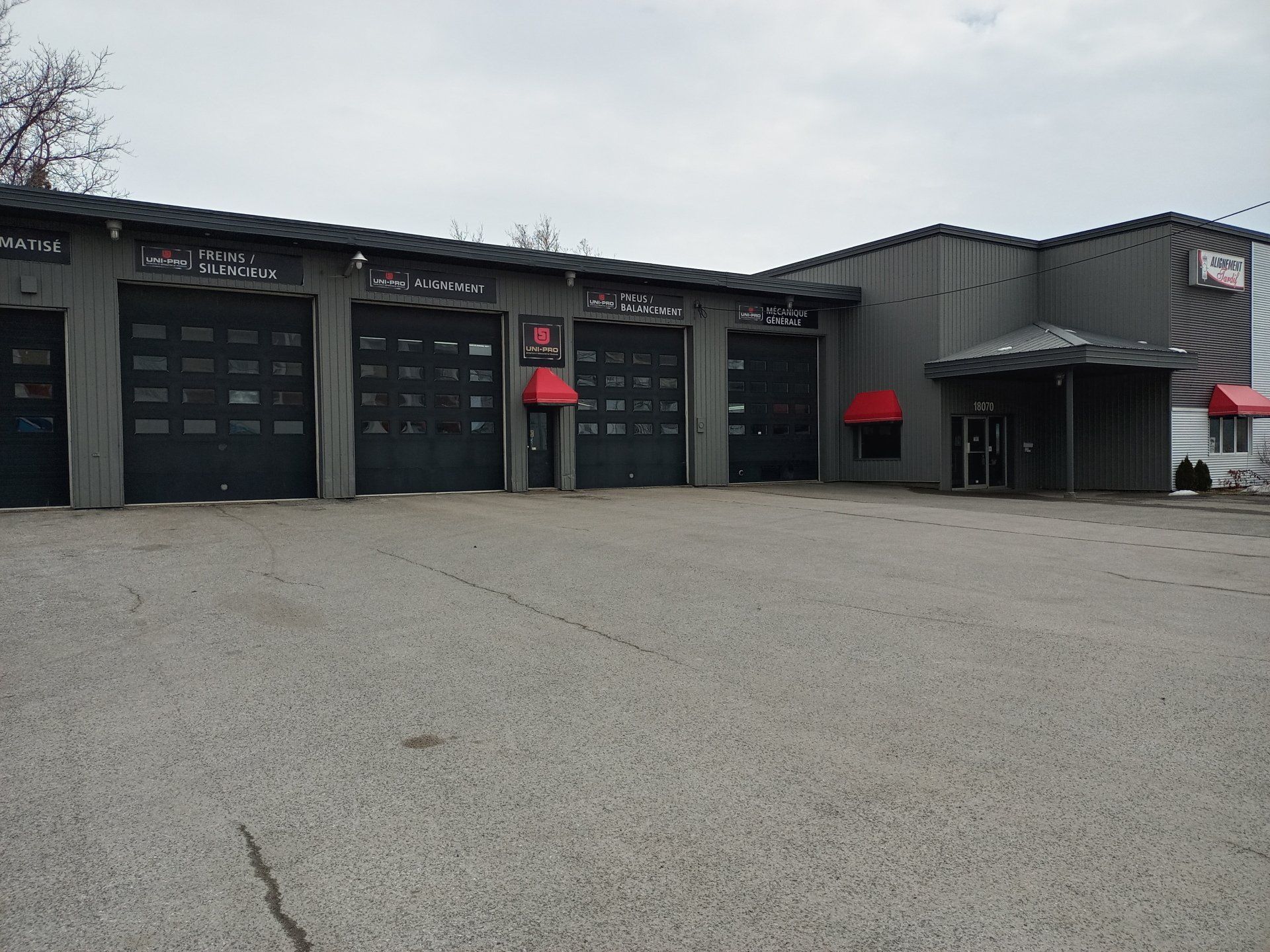Bâtiment industriel gris avec portes de garage noires, auvents rouges et un terrain gravillonné sous un ciel couvert.