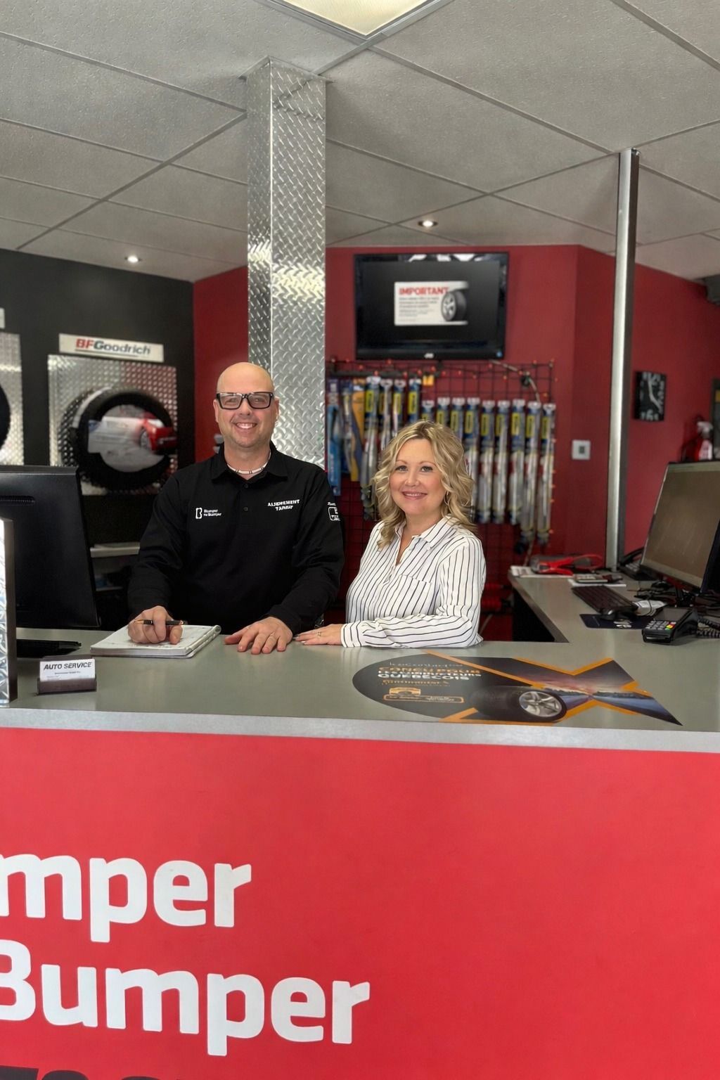 Deux employés se tiennent derrière un comptoir de service rouge dans un magasin, avec des pneus de voiture et des présentoirs derrière eux.