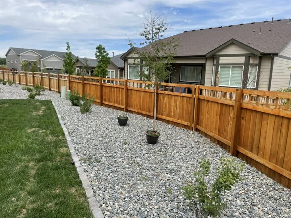 Backyard with a wooden fence, gravel path, small trees, and a house in the background