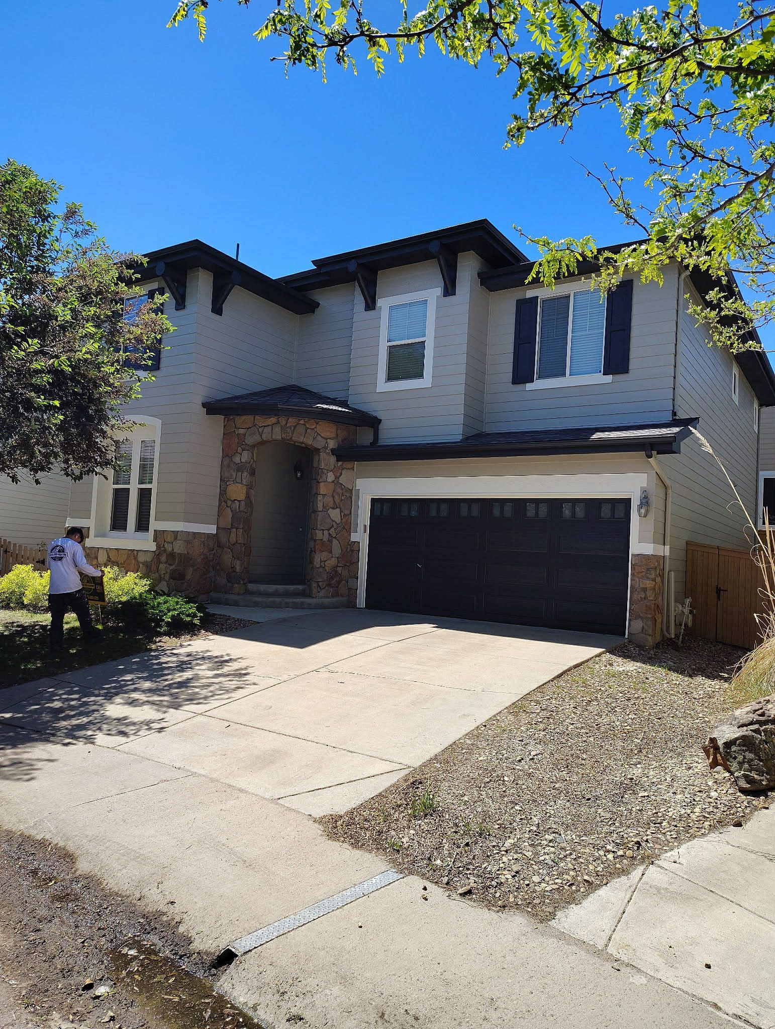 Two-story suburban house with a stone entry, dark garage, and a person in the front yard under a clear blue sky