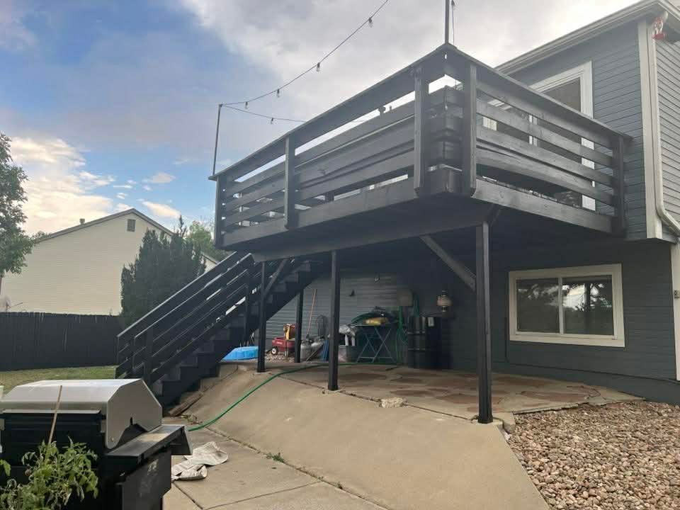 Back deck and staircase on a house with gray siding and black railings, over a concrete patio