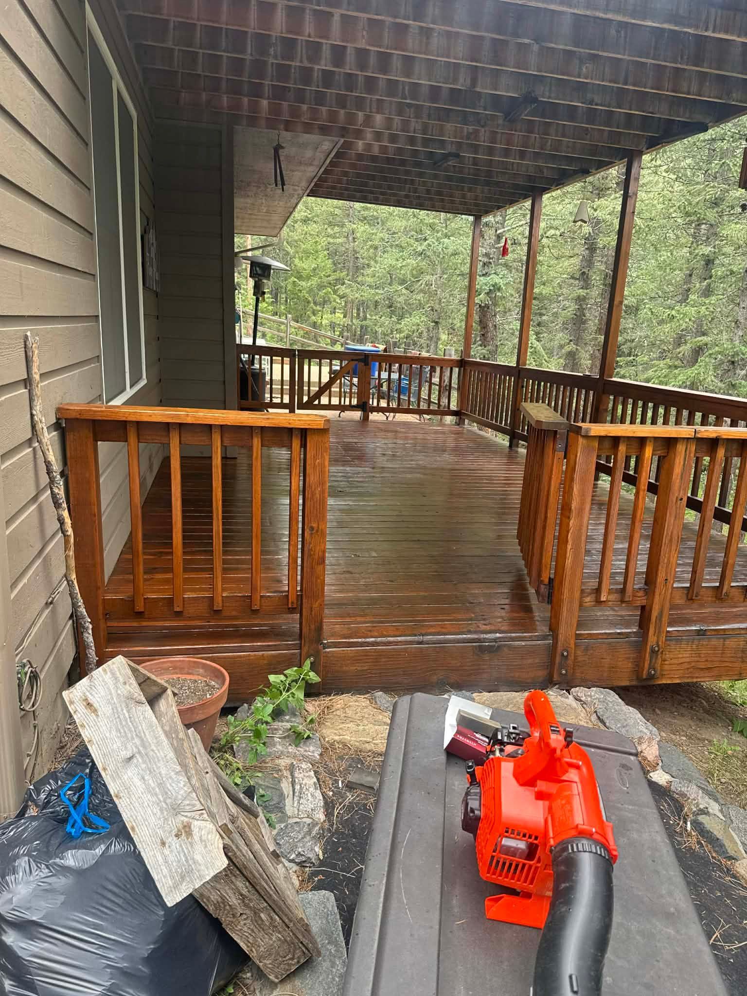 Covered wooden porch with railing and an orange leaf blower on the floor beside a stone and planter