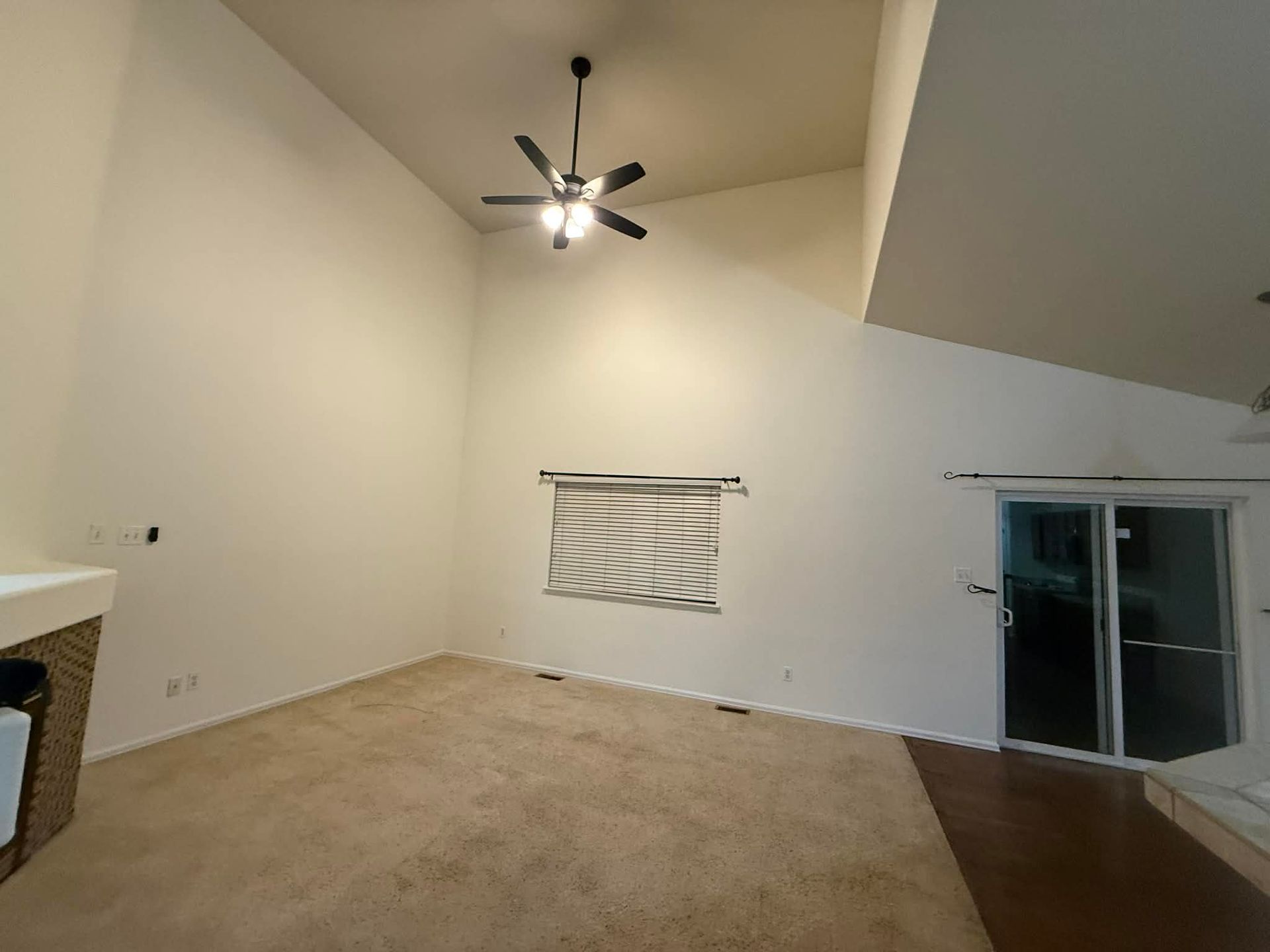 Empty beige living room with high ceiling, ceiling fan, sliding glass door, and a covered wall opening