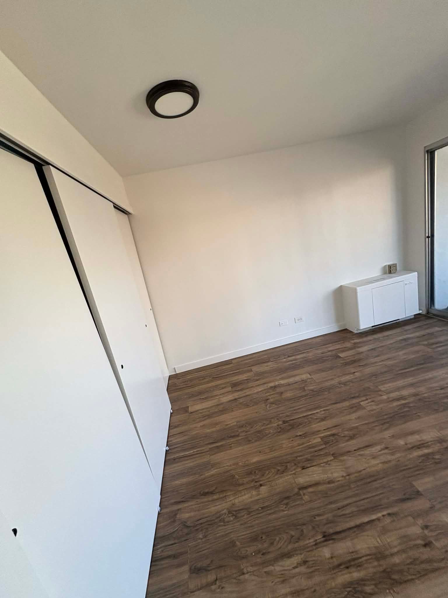 Empty white room with wood flooring, ceiling light, and a radiator near the window.