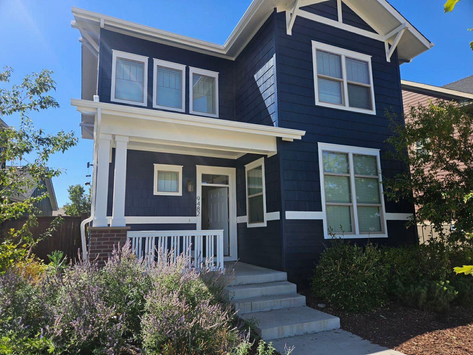 Blue two-story house with white trim, front porch, steps, and shrubs under a clear sky