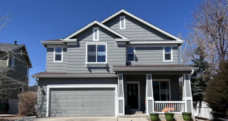 Gray two-story suburban house with white trim, front porch, and two-car garage under a clear blue sky