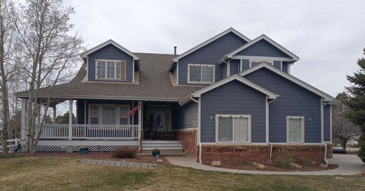 Gray two-story house with a covered front porch and brick foundation, viewed from the street.