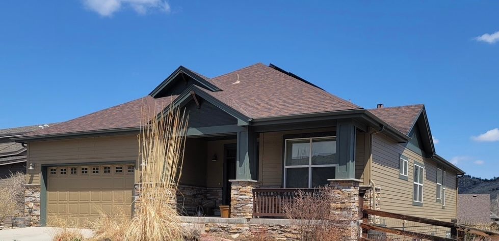Single-story tan house with a brown roof, brick porch, and dry grass in front.