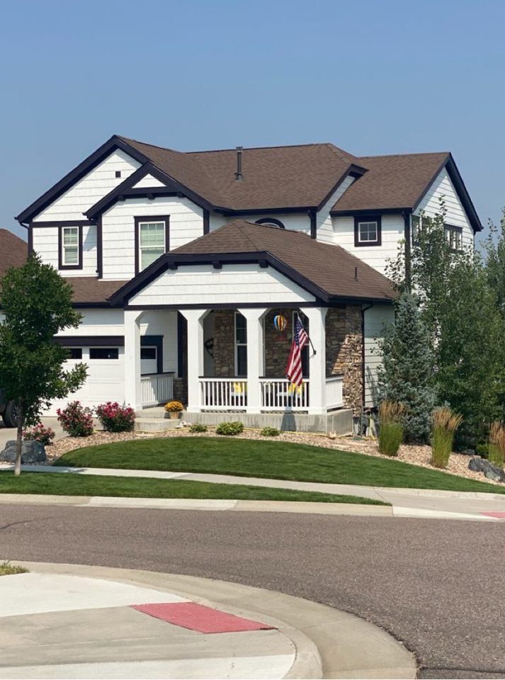Suburban two-story house with white trim, brown roof, front porch, and manicured lawn on a quiet street