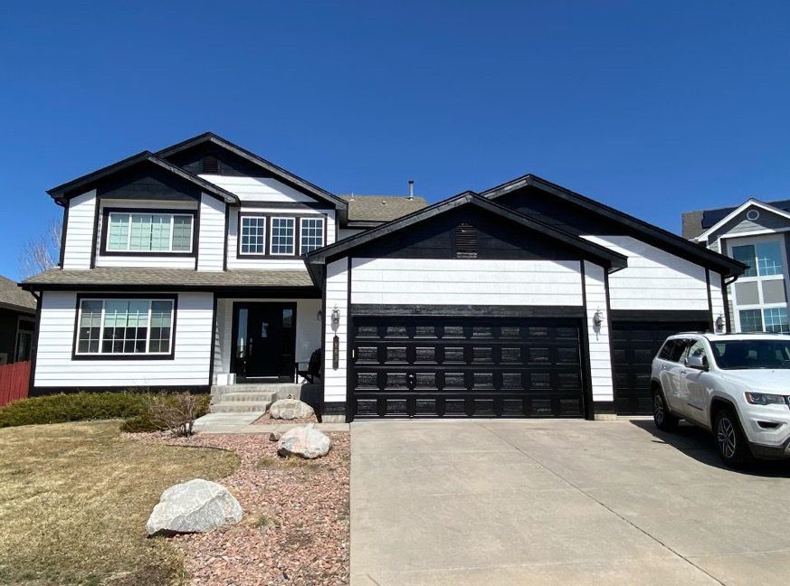 Two-story white house with black trim, black garage doors, driveway, and a parked SUV under a clear blue sky