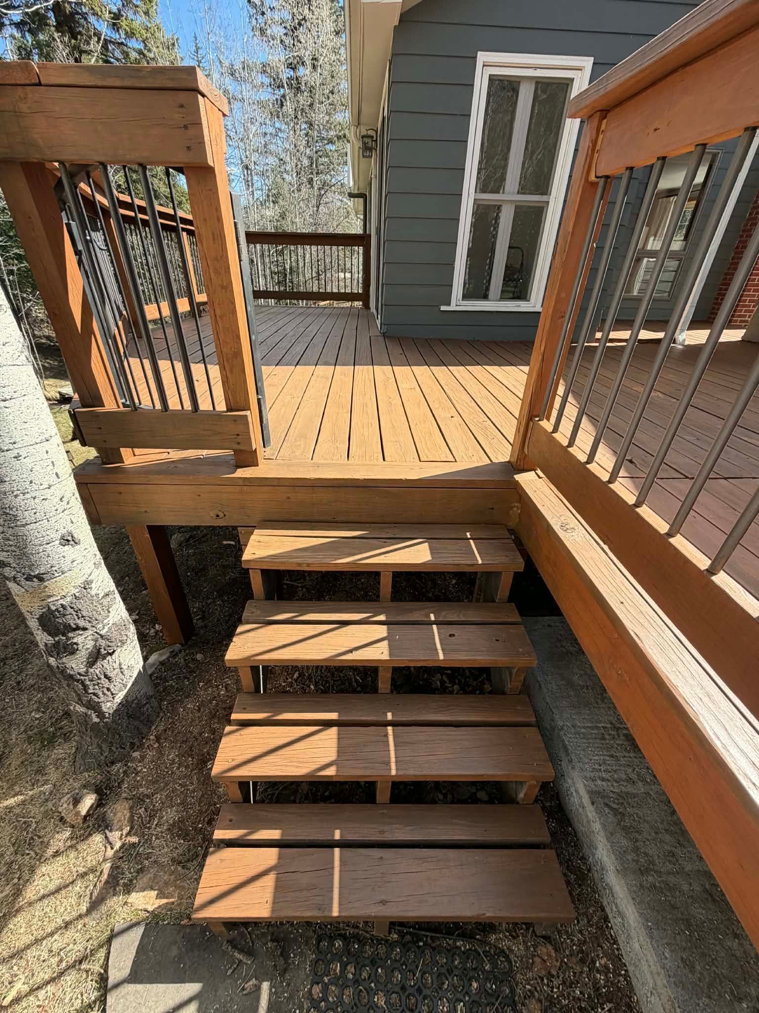 Wooden deck stairs with railings leading to a small landing beside a house and tree trunk