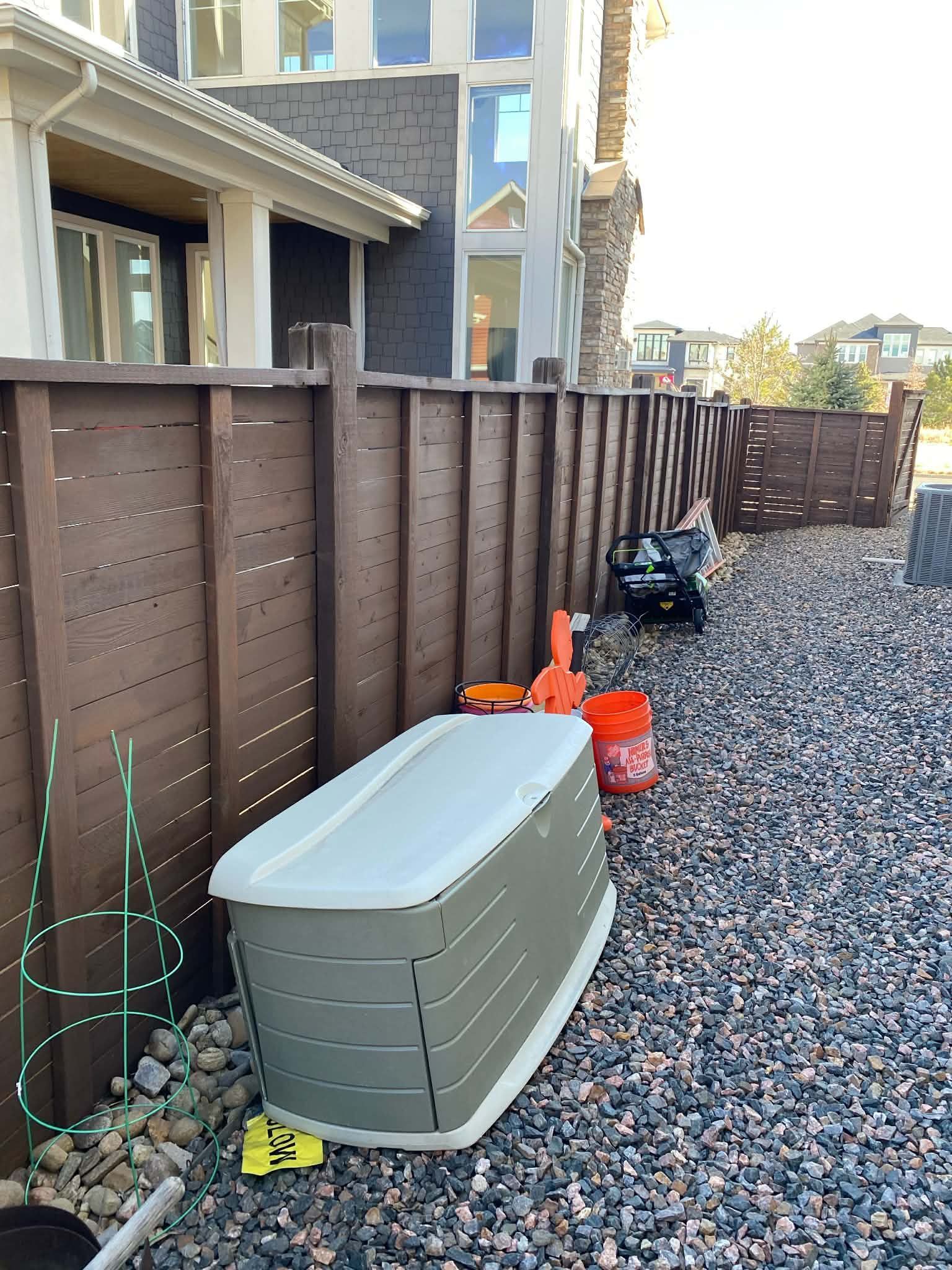 Narrow side yard with brown fence, gray storage box, gravel ground, and orange cones near a house.