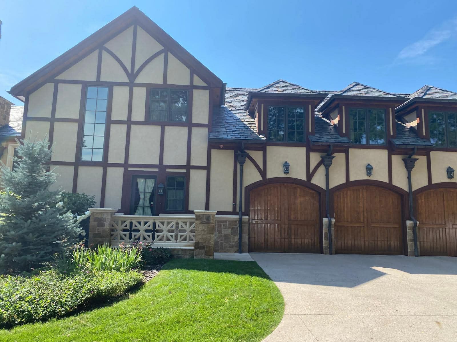 Large Tudor-style house with tan wood trim, arched garage doors, and a green lawn under a bright sky