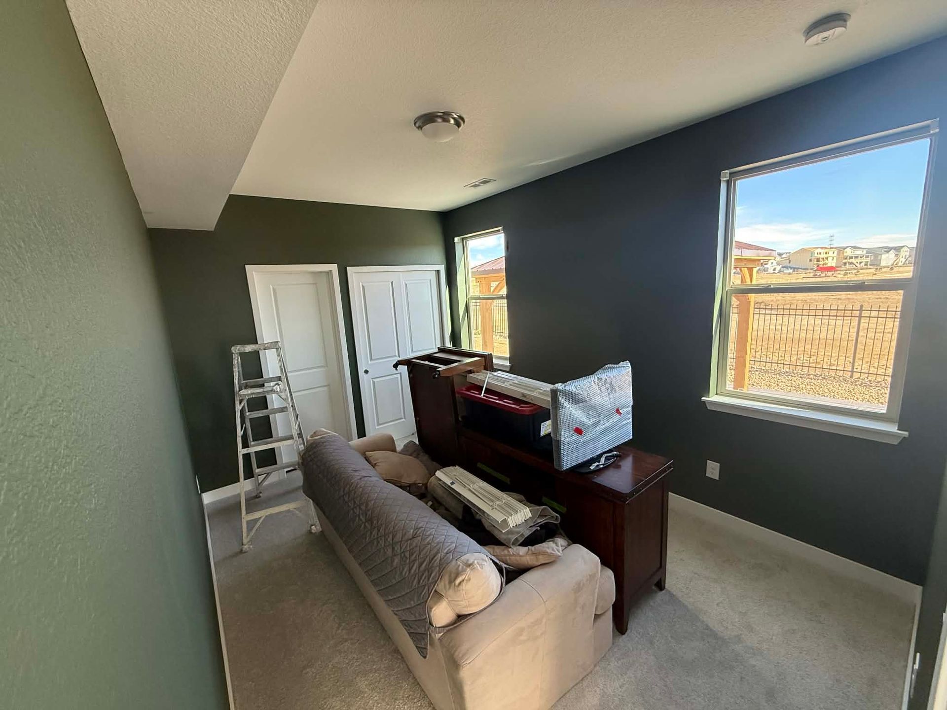 Small room with a gray couch, dresser, ladder, and window, being arranged or cleaned.