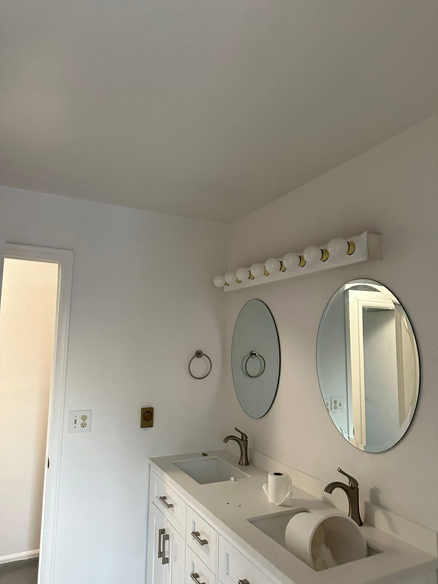 Bright bathroom with double sinks, oval mirrors, and white cabinetry beneath a wall light.