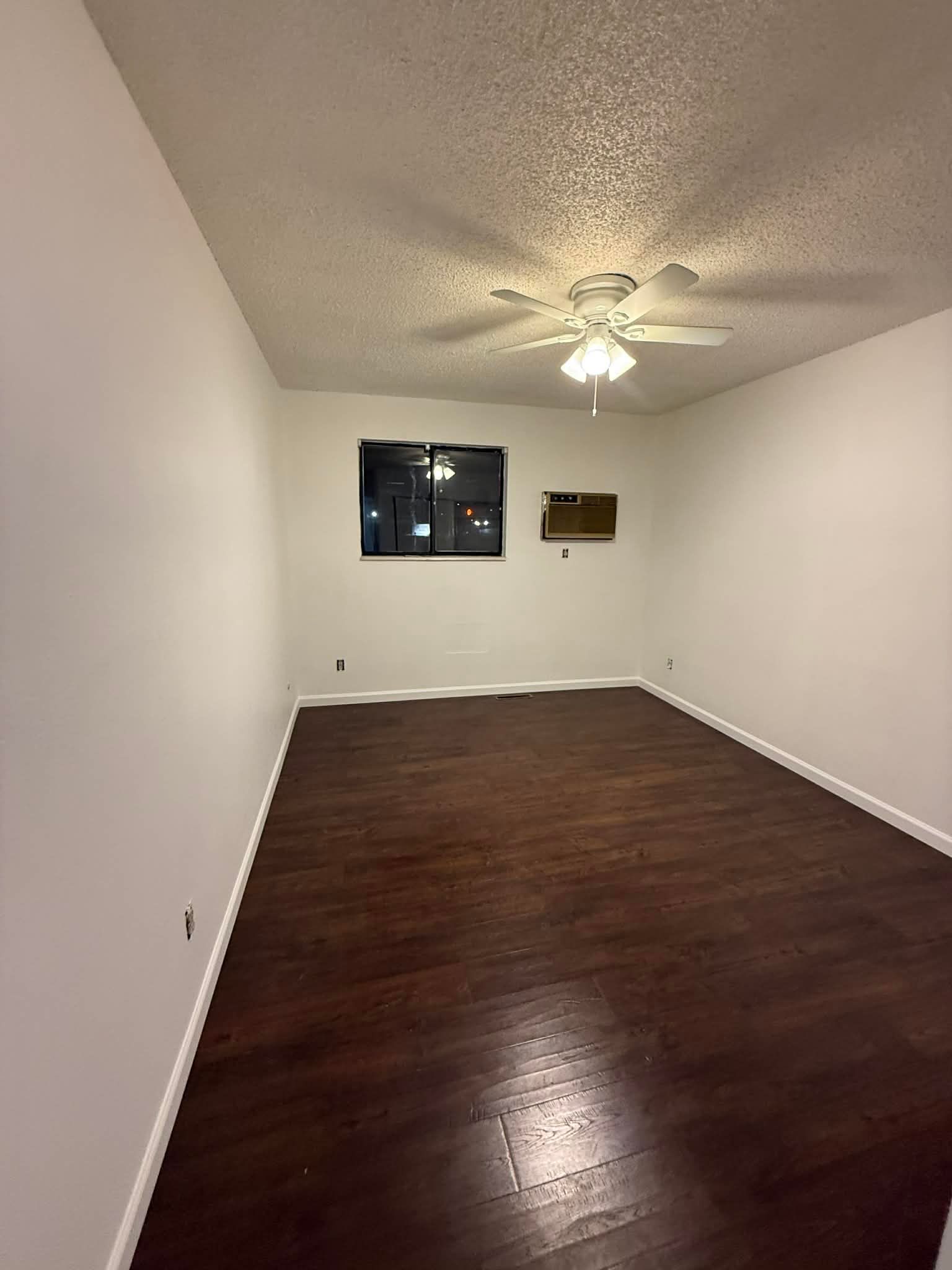 Empty unfurnished room with dark hardwood floors, white walls, ceiling fan, and a window.