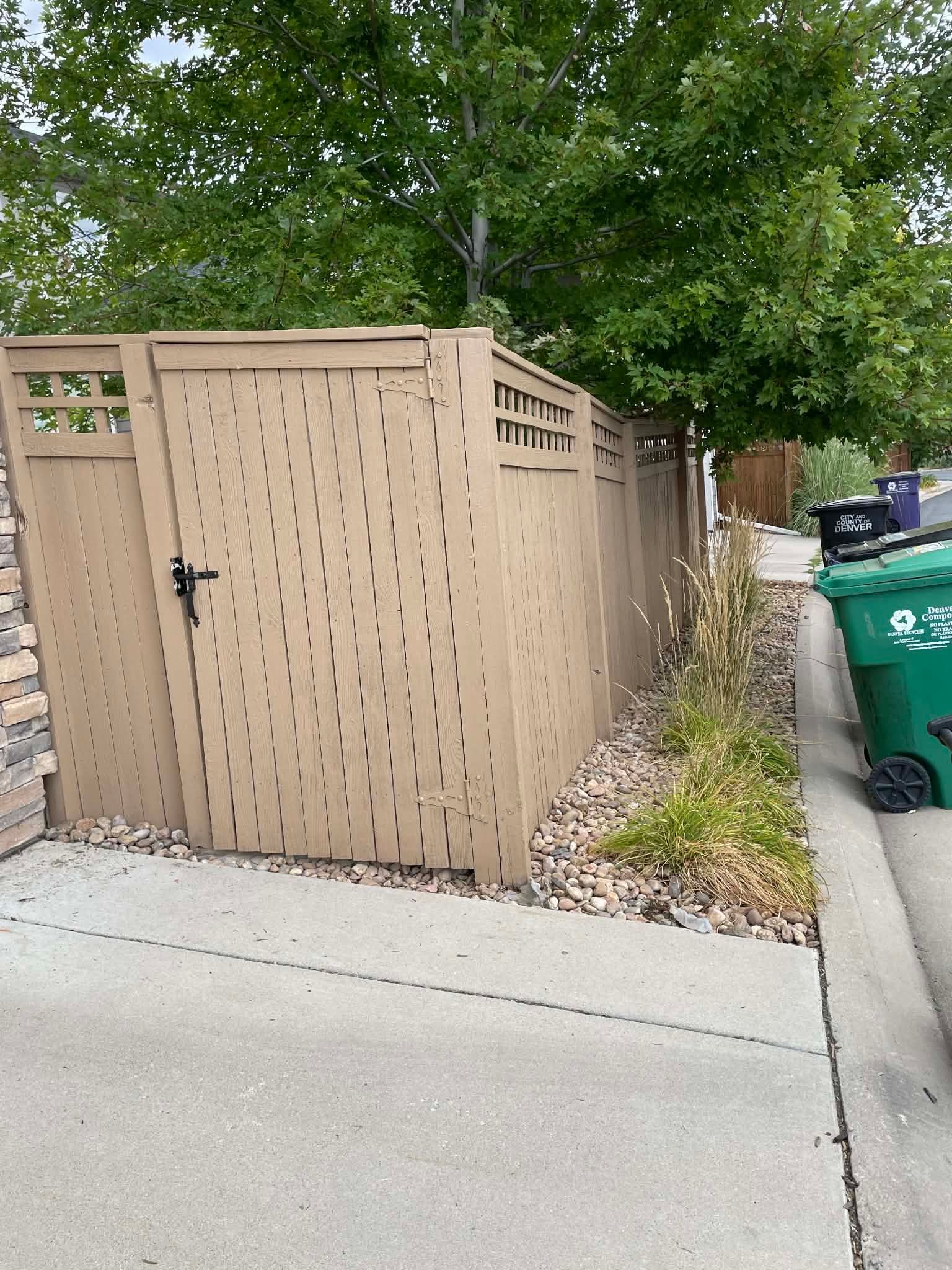 Tan wooden utility enclosure beside a sidewalk, with a large tree and recycling bins nearby.