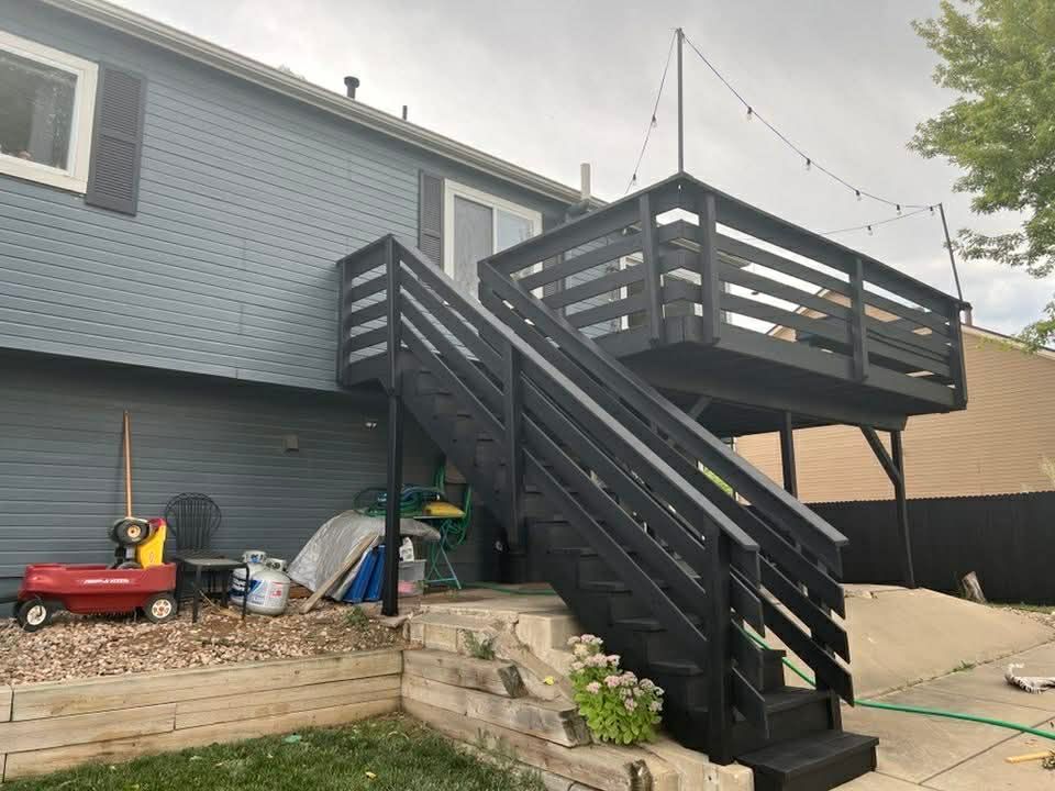 Backyard deck with dark stairs and railing attached to a blue house, with garden tools below