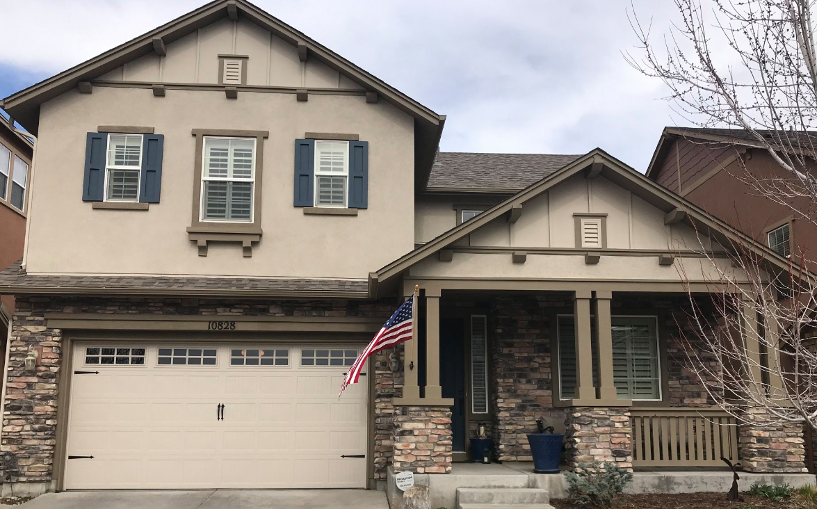 Suburban two-story house with beige siding, stone porch, blue shutters, and an American flag by the garage