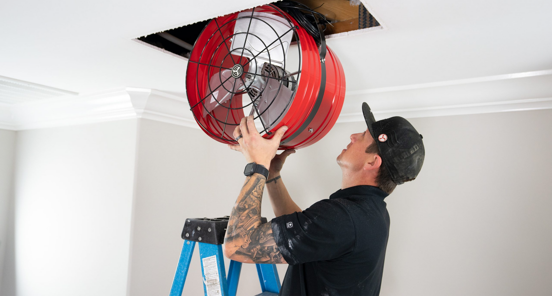 A man is standing on a ladder installing a whole house fan in the ceiling.