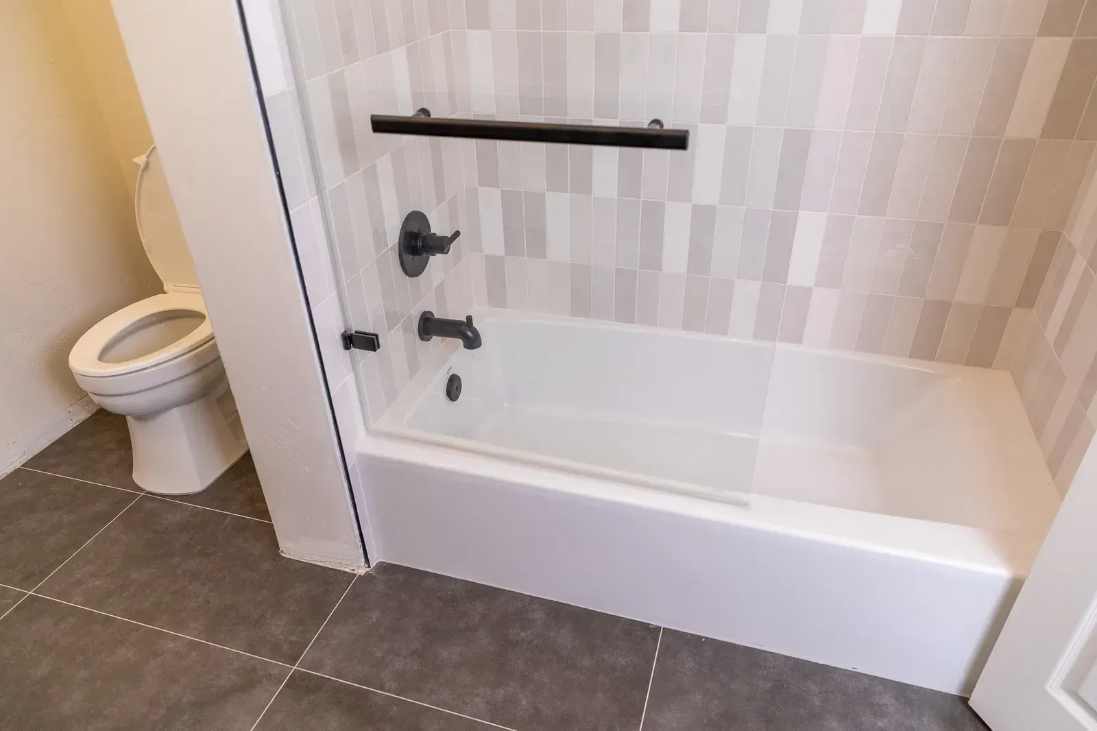 Bathroom with a white tub, black grab bar, tile surround, and toilet beside it.