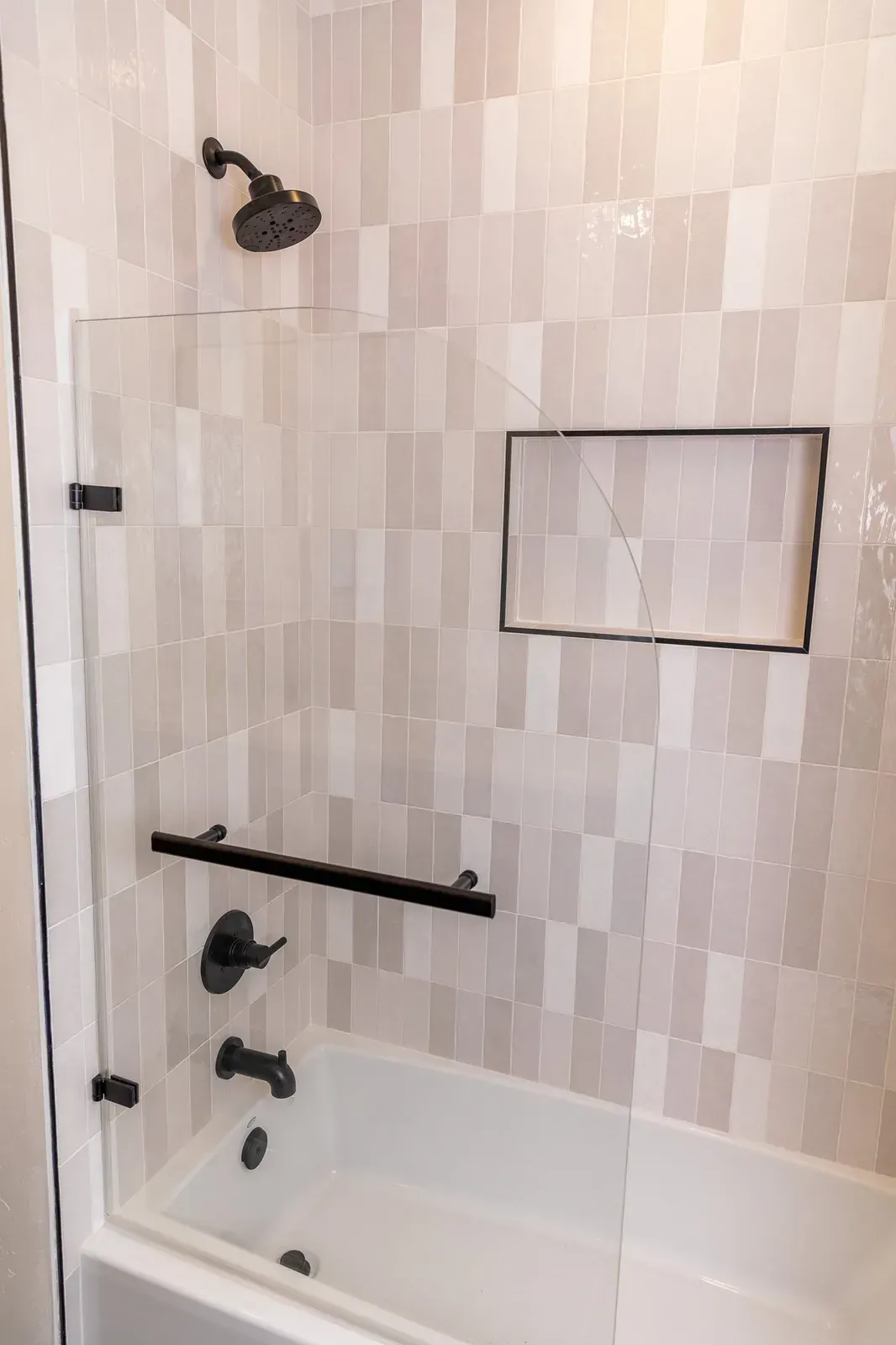 Bathroom with tiled shower wall, wall-mounted showerhead, grab bar, and bathtub below a recessed niche.
