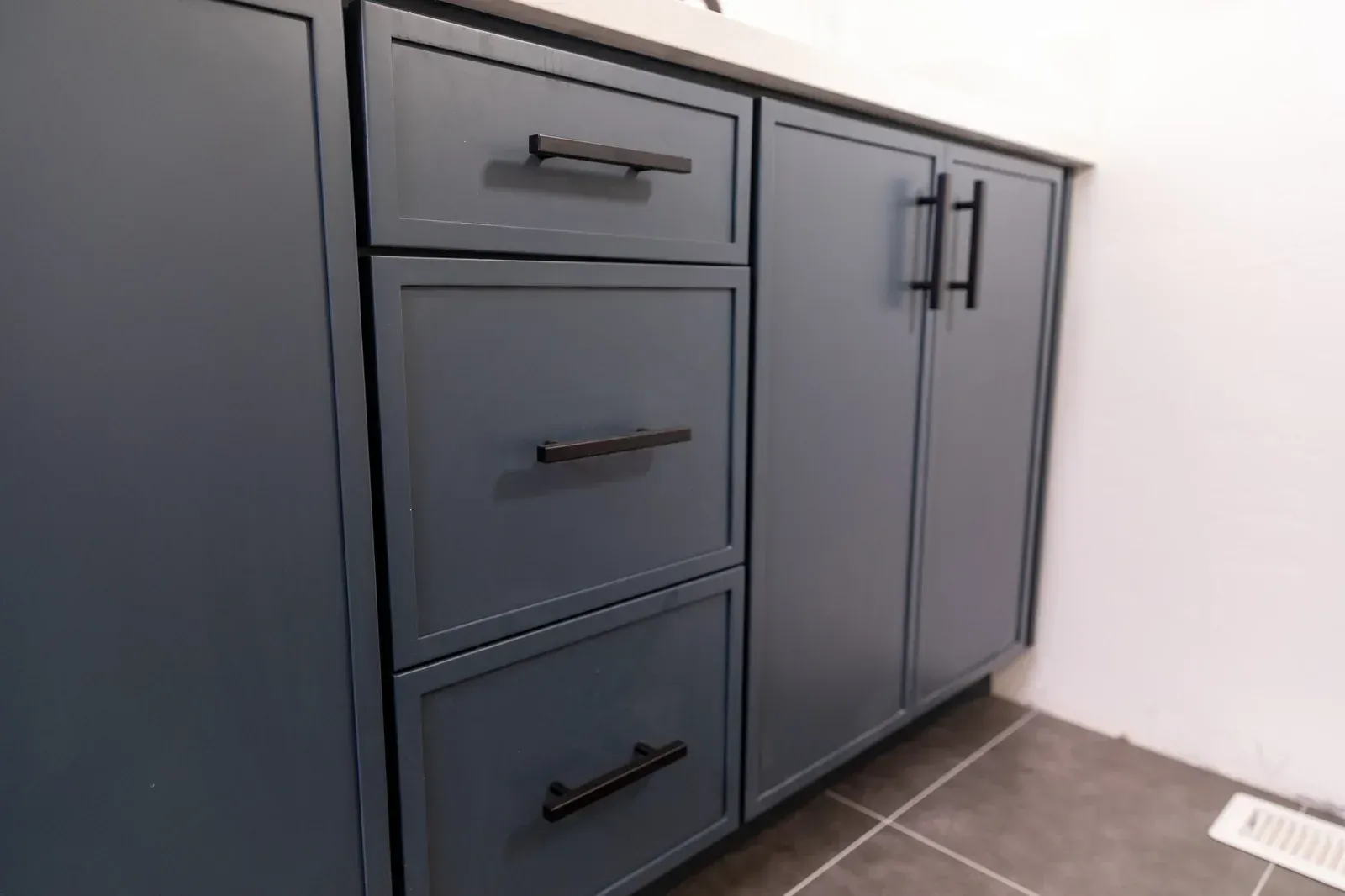 Blue-gray bathroom vanity with drawers and double cabinet doors against a white wall, tiled floor visible