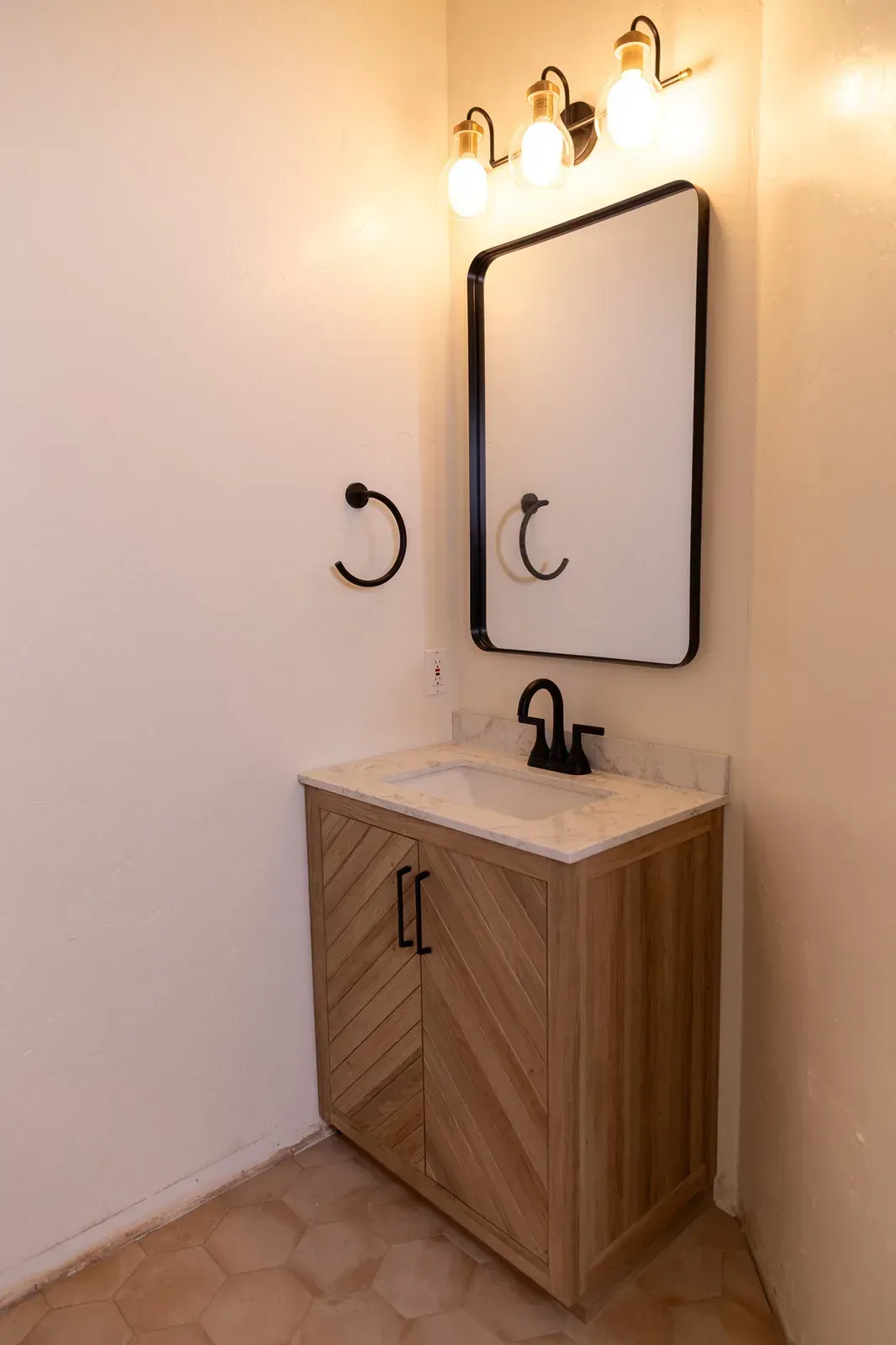 Small bathroom vanity with black faucet, round mirror, and wall sconce lighting on cream walls.