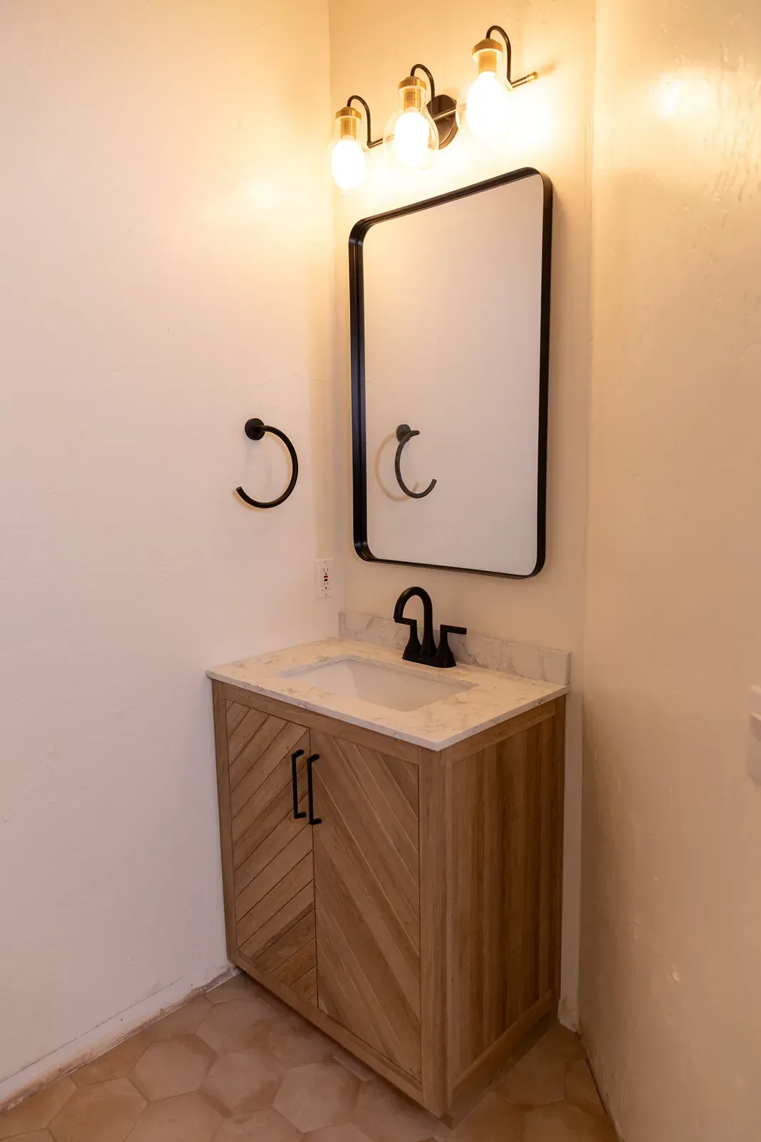 Small bathroom vanity with wood cabinet, white sink, black mirror, and wall sconces.