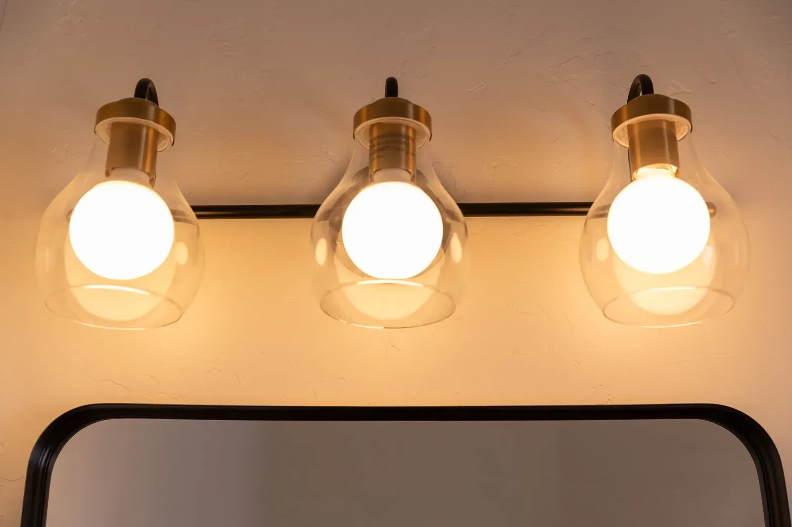 Three lit wall sconces with clear glass shades above a black-framed mirror