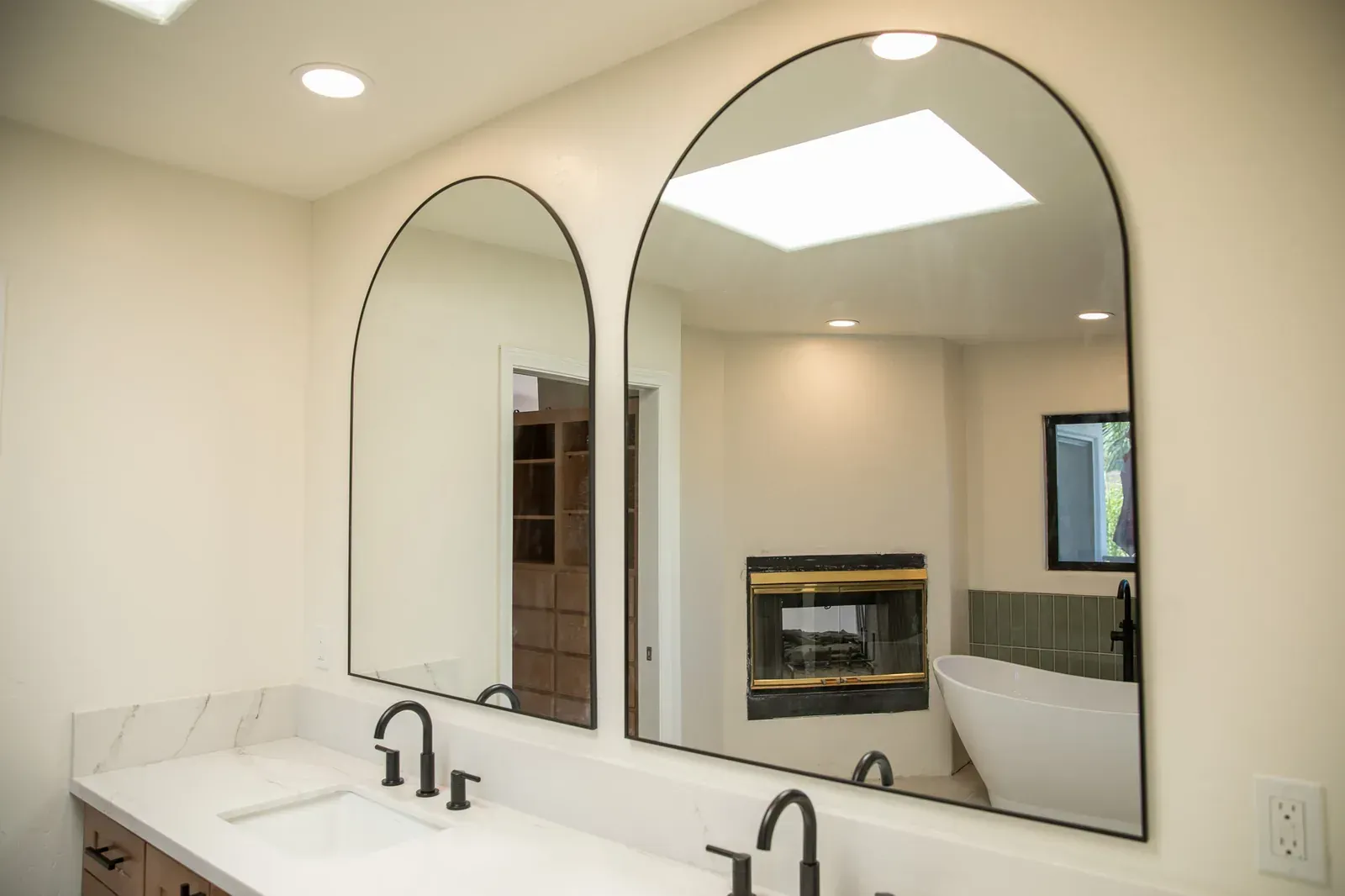 Bathroom vanity with two arched mirrors, white countertop, and a wall-mounted fireplace reflection