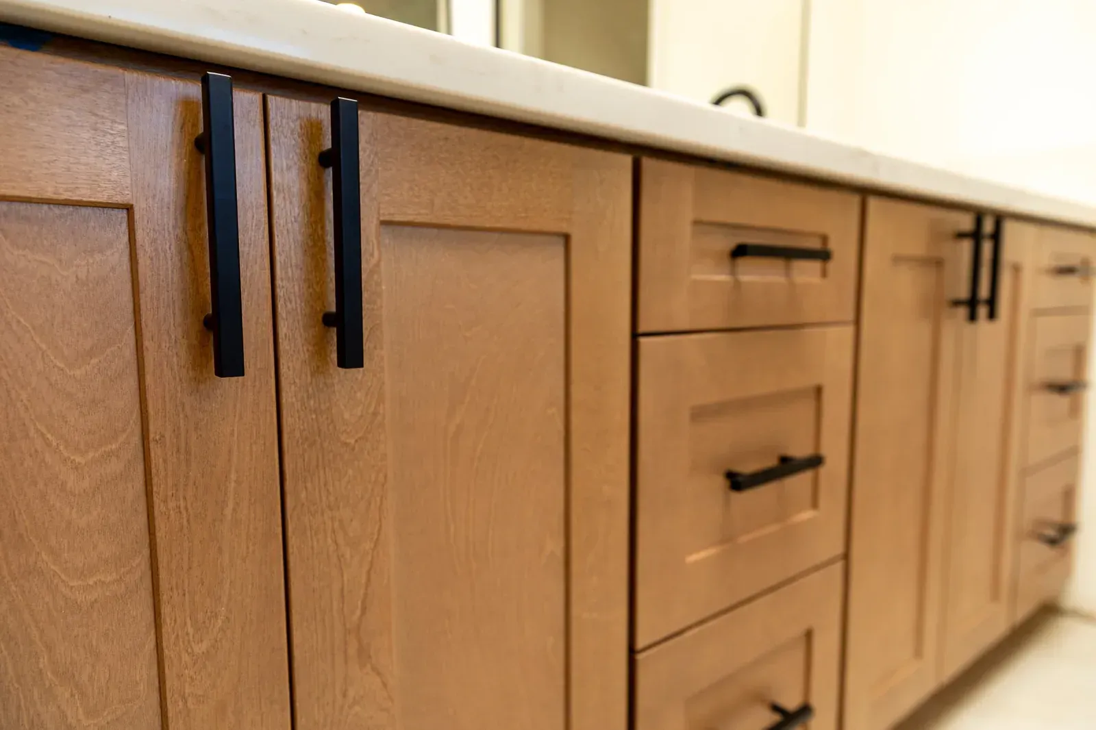 Wooden kitchen cabinets with black pulls beside a light countertop, viewed at an angle.