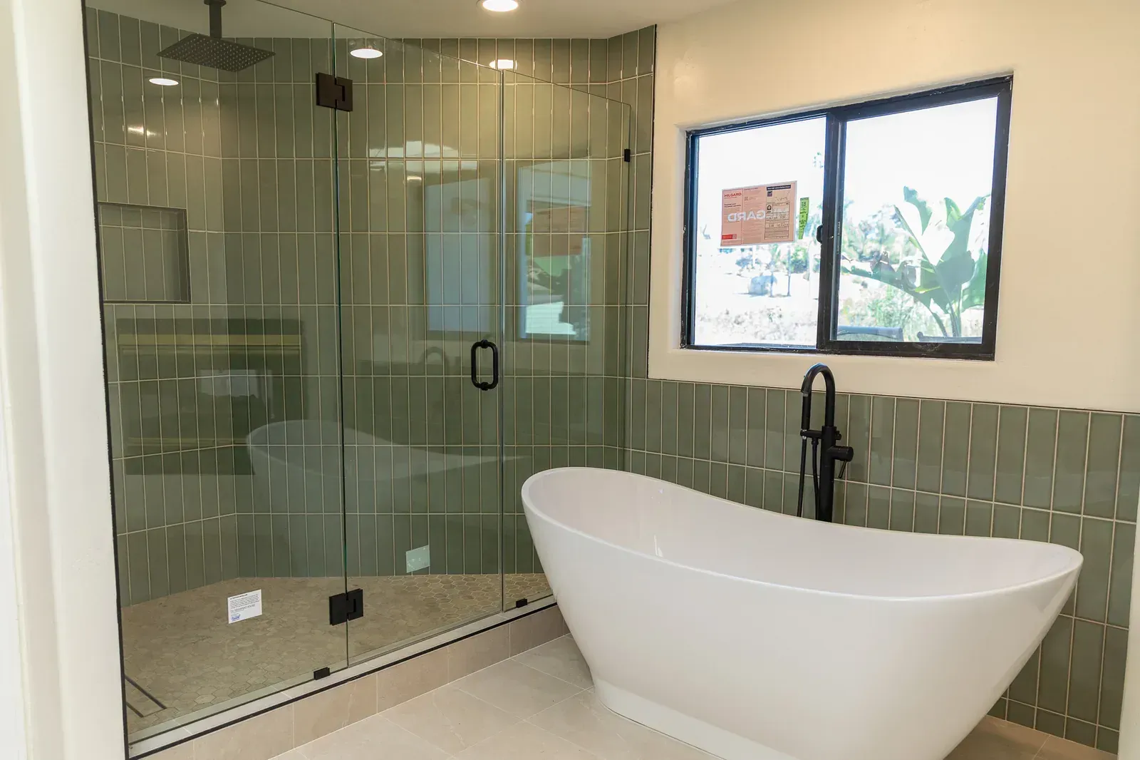 Modern bathroom with glass shower, freestanding tub, green tile walls, and a window.
