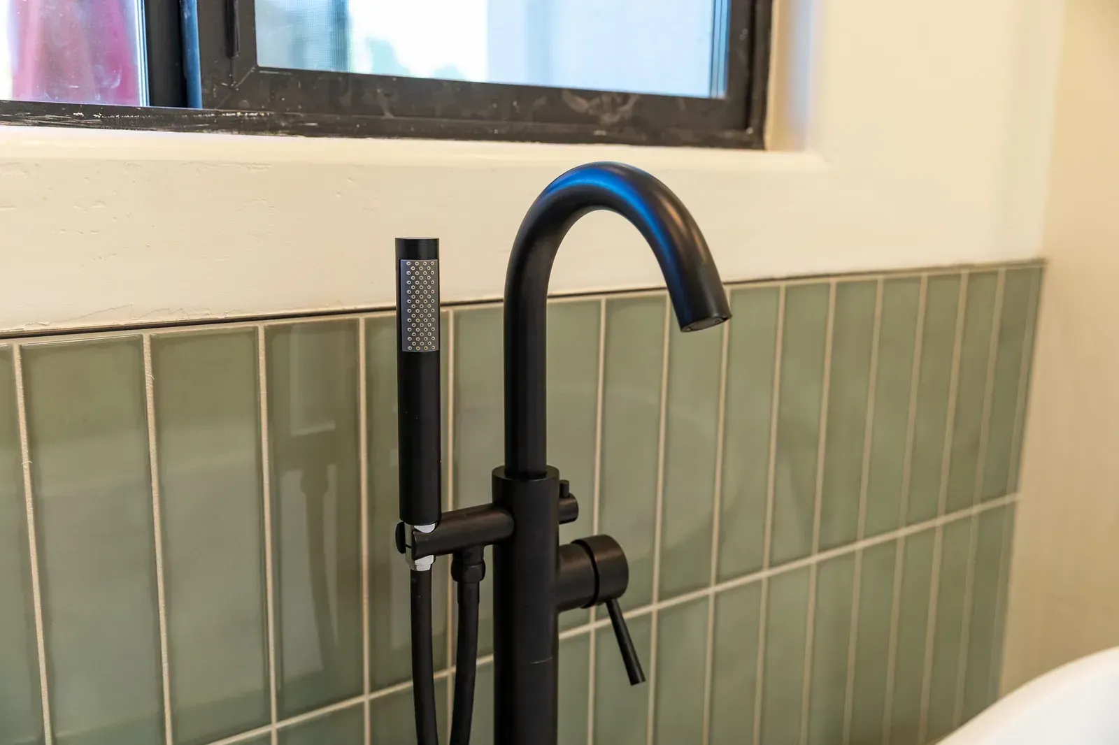 Black matte bathroom faucet with handheld sprayer above green tile backsplash under a window